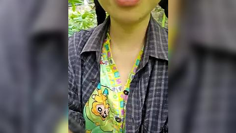 Media: Video of a woman in a green plaid shirt over a yellow and pink patterned top with a cartoon pig graphic. She is standing in a lush, green forest background.