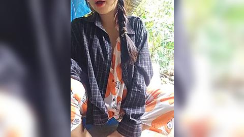 Media: Video of a young woman with medium skin tone, wearing a plaid shirt over a white and orange patterned top, sitting on a beach towel with blurred greenery in the background.