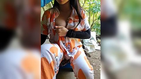 Media: A video shows a woman in an orange and white floral pajama set, sitting outside with a black arm sling. She has long, dark hair and is in a rural setting with greenery and a white bag in the background.