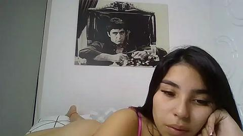 Media: A video of a young woman with straight, dark hair, wearing a pink bra, lying on a bed, looking at a framed black-and-white portrait of a man on a white wall.