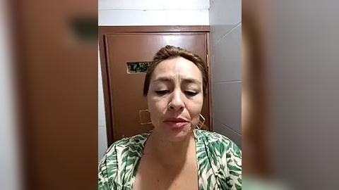 Media: A video of a middle-aged woman with light skin and brown hair, wearing a green floral blouse, appears in a bathroom with white tiled walls and a brown door. She has a neutral expression.