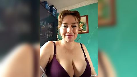 Media: Video of a smiling, middle-aged woman with large breasts, wearing a purple bra, standing in a teal-painted room with framed artwork on the wall.