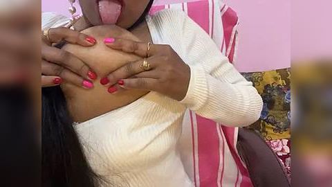 Media: Video of a dark-skinned woman with pink-painted nails, wearing a white sweater, licking her own breast, sitting on a pink and yellow-patterned chair.