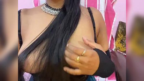 Media: Video of a woman with long black hair, wearing a black strapless top, revealing her breast, and a silver choker necklace, against a pink and white striped background.