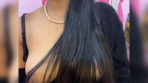 Media: Video of a woman with long, straight black hair, wearing a black off-shoulder top and a pearl necklace, against a blurred pink background.