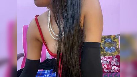 Media: A video of a slender, light-skinned woman with long, straight black hair, wearing a red off-shoulder top, pearl necklace, and black elbow-length gloves, sitting on a pink chair with a colorful cushion in a softly lit room.