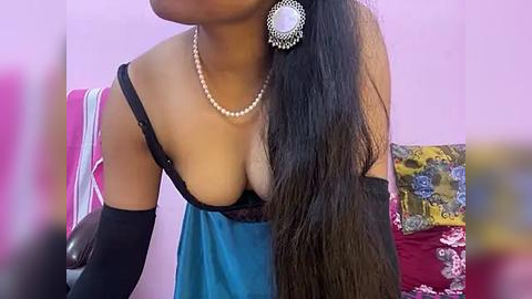 Media: Video of a young woman with medium brown skin and long black hair, wearing a blue and black lingerie top with a pearl necklace and large earrings, seated in a room with pink walls and colorful cushions.