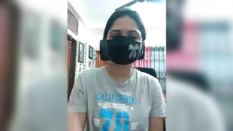 Media: Video of a woman wearing a black face mask and a gray t-shirt with \"California\" in blue, standing in a bookstore with shelves filled with books in the background.