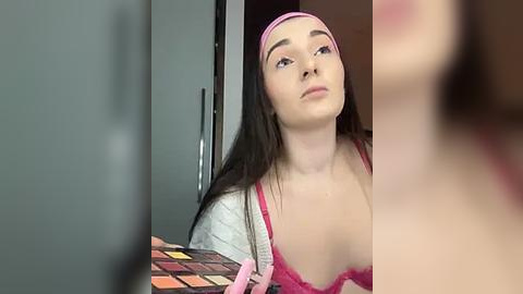 Media: Video of a young woman with long dark hair, fair skin, and pink headband, wearing a pink lace bra, standing in a modern room with grey cabinets.