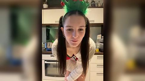 Media: Video of a young woman with straight brown hair in pigtails, wearing a red and white striped shirt and a green elf hat, standing in a modern kitchen with white cabinets and a microwave.