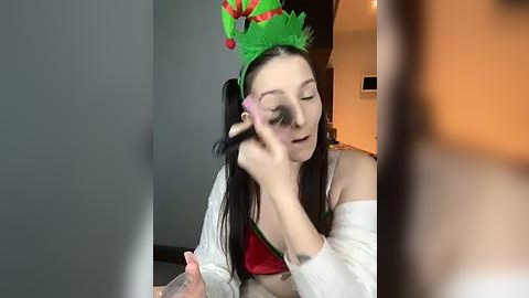 Media: Video of a young woman with pale skin and long black hair, wearing a festive green elf hat, red bra, and white cardigan, applying makeup with a black brush. Background shows a dimly lit room with blurred furniture.