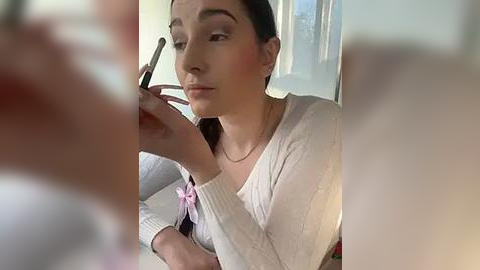 Media: A video of a woman applying makeup with a brush, wearing a white cardigan, against a blurred background.