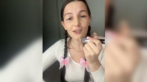 Media: Video of a young woman with fair skin, brown hair, and light makeup applying foundation with a brush. She wears a white shirt with pink bows and a black cardigan. Background is blurred.