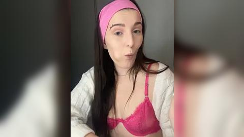 Media: Video of a young woman with fair skin and long brown hair, wearing a pink headband, pink lace bra, and white cardigan, making a pouty face.