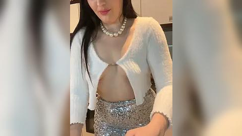 Media: A video of an Asian woman with long black hair, wearing a white, open-front cropped sweater and a silver sequined skirt, sitting indoors.