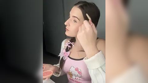 Media: Video of a young girl with fair skin and long brown hair, wearing a pink Hello Kitty shirt, applying makeup. She holds a pink brush and a compact mirror.