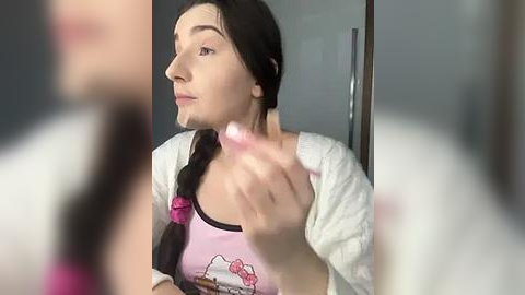 Media: Video of a woman with long black hair in braids, wearing a pink tank top and white cardigan, applying lipstick in a mirror.