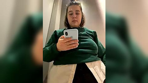 Media: Video of a plus-sized woman with medium-length brown hair, wearing a green long-sleeve shirt, white jacket, and black pants, taking a selfie in a bathroom mirror.