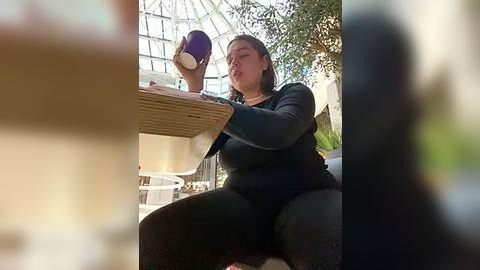 Media: Video of a plus-size woman with short dark hair, wearing a black sweater and jeans, drinking from a purple coffee cup, seated at a wooden table in a sunlit, glass-roofed indoor caf\u00e9 with greenery in the background.