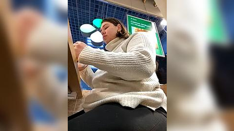 Media: Video of a woman in a cream sweater and black pants, sitting and holding a tablet, in a modern, tiled setting with a green sign.