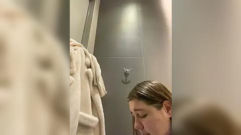 Media: A video of a bathroom showing a woman with light skin, brown hair, and glasses, holding a beige towel, standing near a shower door with a metal hook.