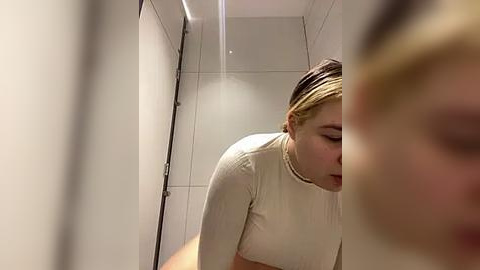 Media: Video of a young woman with light skin, short blonde hair, wearing a beige sweater, looking down in a tiled bathroom with a reflective mirror.