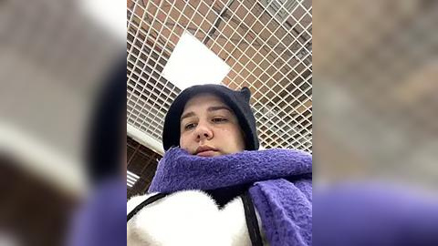 Media: A video of a person wearing a black cat hood and purple blanket, standing in a room with a white ceiling and grid pattern. The image is slightly out of focus.