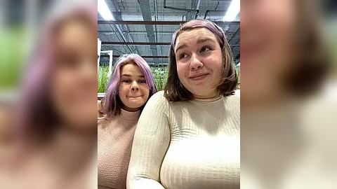 Media: Video of two women with shoulder-length hair, one with purple, the other with brown, wearing light-colored sweaters, standing in a greenhouse with plants in the background.