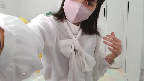 Media: Video of an Asian woman with straight black hair, wearing a light pink face mask and white lab coat, taking a selfie in a white-walled room with a yellow object and closed white cabinets in the background.