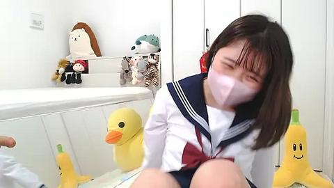 Media: Video of a young girl with straight black hair wearing a white and navy school uniform, seated on a bed with plush toys, including a duck and bear.