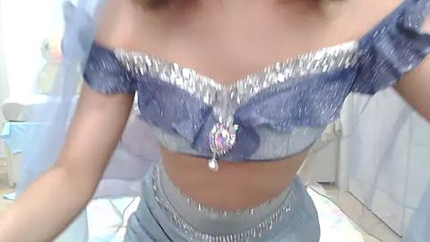 Media: A video of a woman in a sparkling, off-the-shoulder blue genie costume with a jeweled midriff, seated on a colorful quilted surface, in a softly lit room with pastel decor.