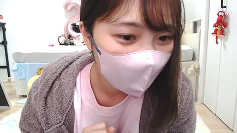 Media: Video of an East Asian woman in a pastel pink mask, gray cardigan, and pink shirt, sitting on a white floor, surrounded by plush toys in a bright, clean bedroom.