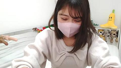 Media: A video of a young Asian woman in a pink face mask, wearing a white robe, sitting in a brightly lit room with a bed, colorful toys, and a yellow plush toy in the background.