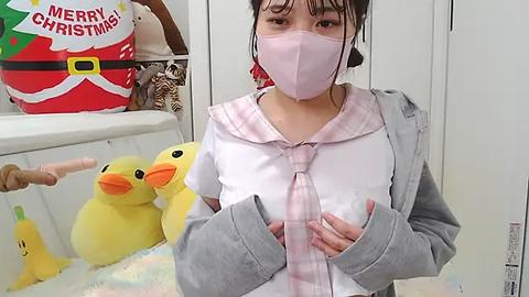 Media: Video of a young Asian woman with light skin, wearing a pink face mask, a pink and white plaid shirt, and a grey hoodie, holding a plush yellow duck, in a festive room with Christmas decorations.