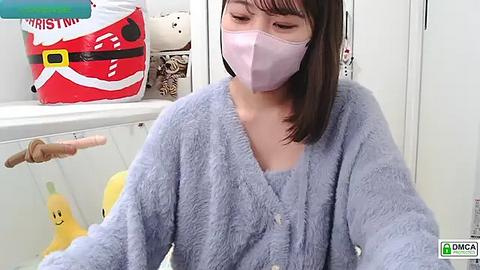 Media: Video of an East Asian woman with shoulder-length dark hair, wearing a light blue fleece robe, a pink face mask, and a brown belt, seated in a white room with festive Christmas decorations and a stuffed toy.