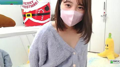 Media: A video of an Asian woman with shoulder-length brown hair, wearing a light blue fuzzy cardigan, pink mask, and white gloves, sitting on a white floor. Background includes a red and white Christmas box, a yellow banana toy, and a blue pillow.