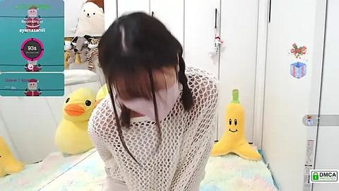 Media: Video of a young Asian woman with long black hair, wearing a white mesh top, bending over in a pastel-colored room with plush toys, including a yellow duck and a smiling carrot.