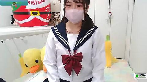 Media: Video of an Asian woman in a white school uniform with a red bow, wearing a face mask, standing in a colorful room with stuffed toys and a Christmas bag.