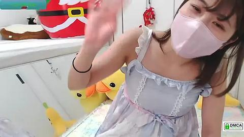 Media: Video of a young Asian woman in a light blue dress, pink face mask, and white mask. She is in a playful, childlike setting with plush toys and a festive background.