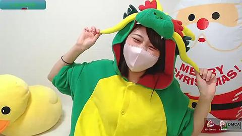 Media: Video of a person in a green and yellow dragon onesie, with a white face mask, playfully adjusting their hood. Background includes a Santa Claus decoration and a yellow duck toy.