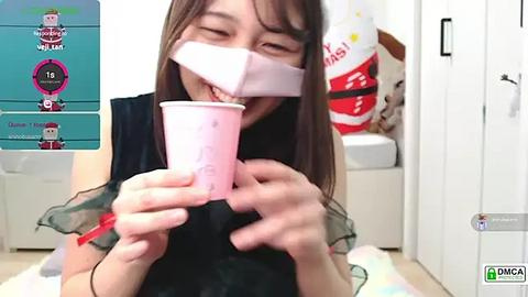 Media: A video of an Asian woman with long brown hair, wearing a black dress, with a pink bandana over her mouth, holding a pink paper cup. Background includes a white door and a colorful banner.