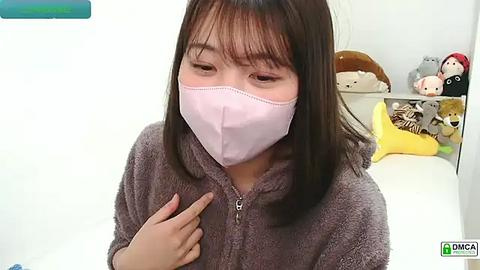 Media: Video of a young Asian girl with straight black hair and light skin, wearing a pink face mask, gray fuzzy robe, and pointing at her chest. Background features stuffed animals, white walls, and a blue piece of furniture.