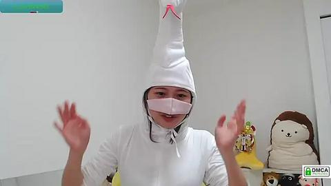 Media: Video of a smiling woman in a white nurse's hat, mask, and uniform, hands raised, in a brightly lit room with plush toys.