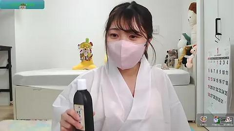 Media: Video of a young Asian woman in a white kimono and pink mask, holding a black bottle in a modern, clean bathroom with a white bathtub, toys, and calendar.