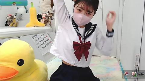 Media: Video of an Asian woman in a schoolgirl outfit, mask, and goggles, playfully posing with a stuffed yellow duck and a stuffed black cat.