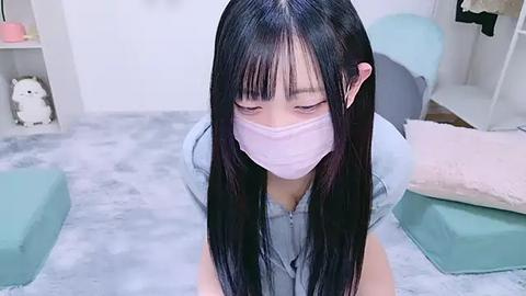 Media: Video of an Asian woman with long black hair, wearing a light blue shirt and a pink face mask, kneeling on a light gray carpet in a pastel-themed room with light blue furniture and a white wall.