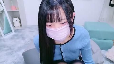 Media: Video of an East Asian woman with long black hair and fair skin, wearing a light blue sweater and pink surgical mask, kneeling on a white carpeted floor in a room with a teal cushion.