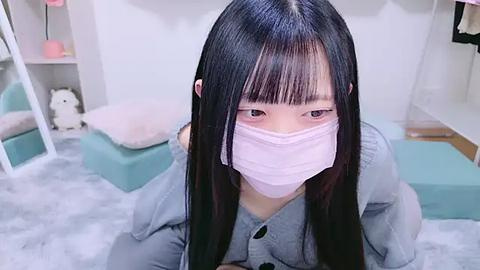 Media: Video of a young Asian woman with long black hair and pale skin wearing a light blue button-up shirt and a pink face mask, kneeling on a white fluffy carpet in a pastel-colored room with a bed and shelves.