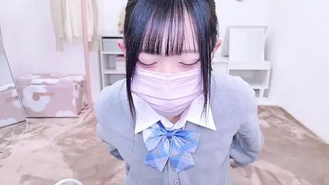 Media: Video of an Asian schoolgirl with straight black hair, wearing a light blue plaid shirt with a bow tie, a white face mask, and a gray cardigan, standing in a minimalist room with a beige carpet and white furniture.