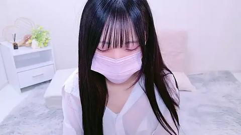 Media: Video of a young Asian woman with long black hair, wearing a white mask, sitting on a white cushion in a minimalist room with a white desk and plants.